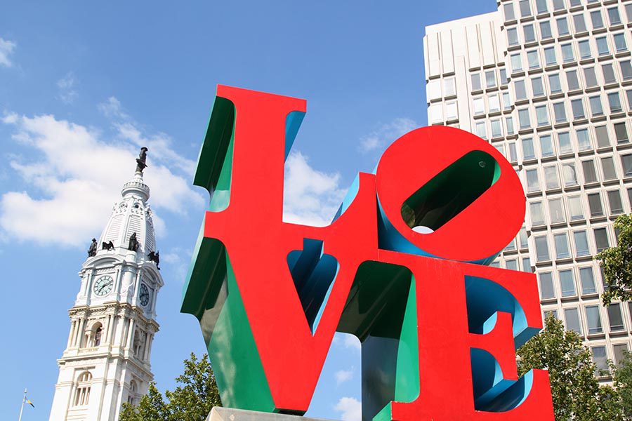 About Our Agency - Statue of the Word Love in a Tree Filled Park in Philadelphia, Blue Sky Overhead and White City Buildings in the Background