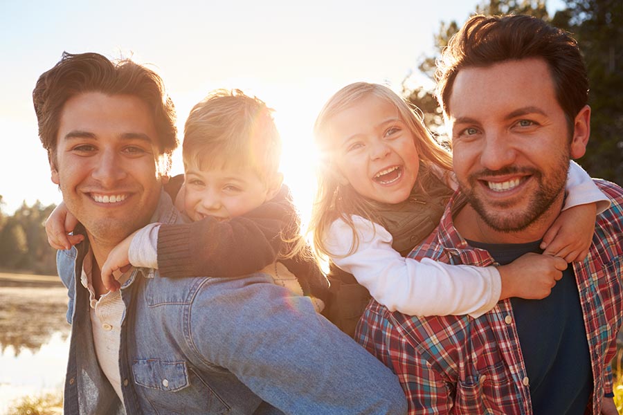 Personal Insurance - Young Family, Kids Riding on Their Dads' Backs, Smiling and Laughing With the Sun Shining Behind Them