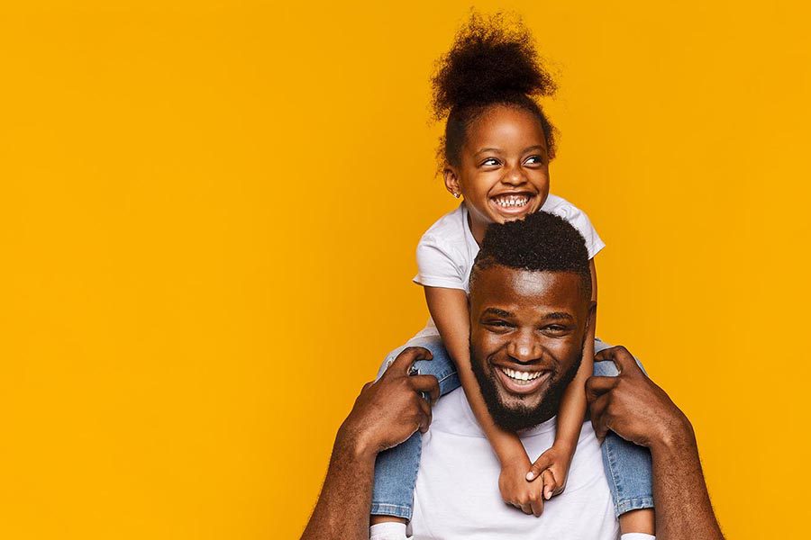 Get a Quote - Young Daughter Sits on Her Father's Shoulders, Both Smiling and Wearing White Shirts, Against a Deep Yellow Background